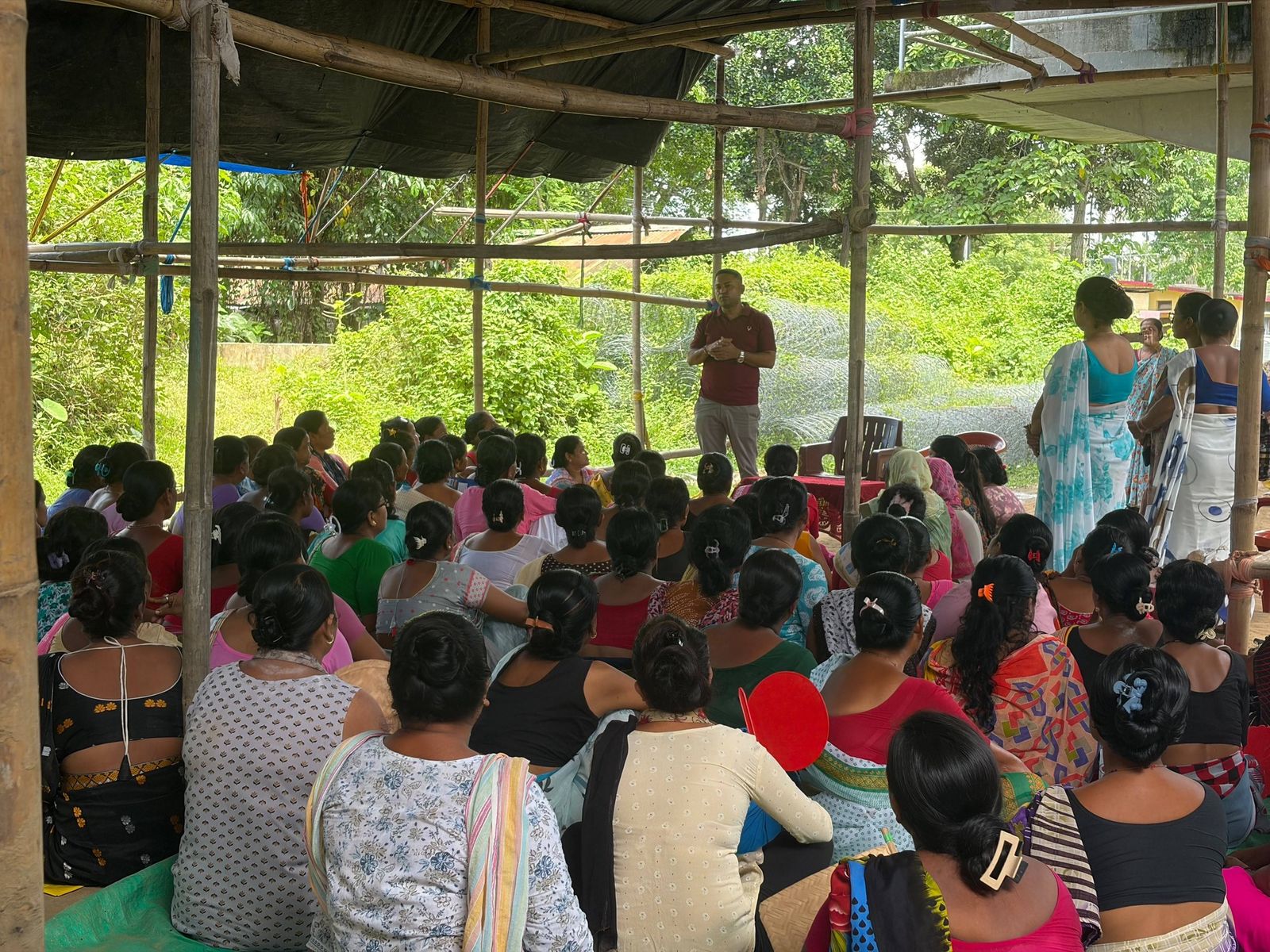 A large group of women seated under a bamboo shed in a green village, listening to a PSF facilitator