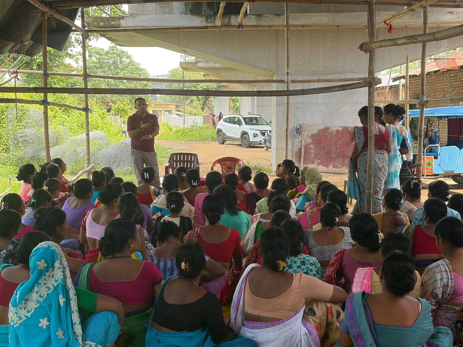 Women seated outdoors at a village meeting while a facilitator in a maroon shirt addresses them